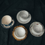 Overhead view of four vintage mismatched teacup and saucer sets arranged on linen, each with unique colors, florals, and gilt details.
