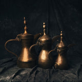 Three vintage brass Turkish coffee pots arranged in soft light, featuring aged patina, tall spouts, and elegant finials.
