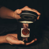 Close-up mobile image of the Elixir of Life candle label and amber glass, focusing on the fine print and rich texture of the vessel.